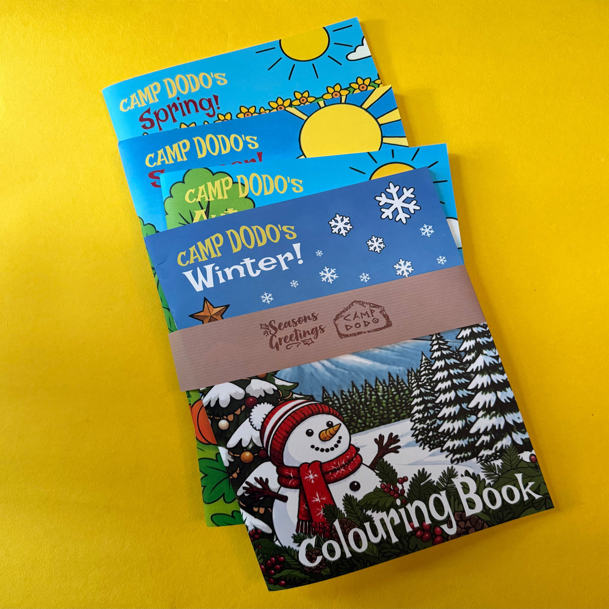 Four  illustrated colouring books fanned out on a bright yellow background. The visible titles are Camp Dodo's Spring,,Camp Dodo's Summer, Camp Dodo's Autumn and Camp Dodo's Winter, with the front book showing a snowy winter scene and the text 'Colouring Book' and 'Seasons greetings from Camp Dodo'.